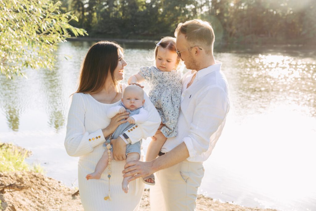 Eine junge Familie aus Vater und Mutter, halten ihre beiden Kinder, ein Kleinkind und Baby, auf dem Arm und lächeln sich an.